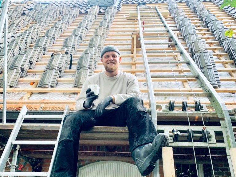 Handwerker sitzt auf einer Leiter vor einem Gerüst und hält ein Werkzeug in der Hand.