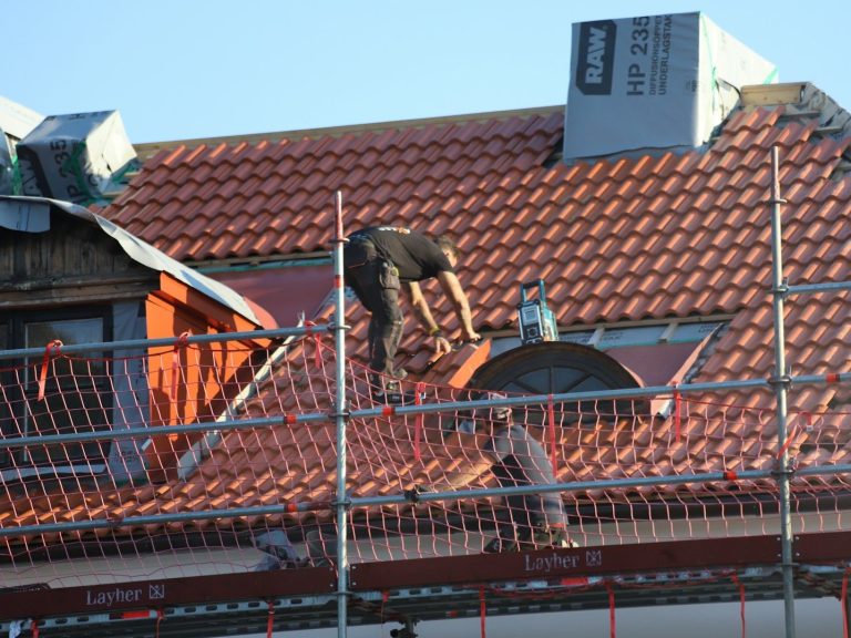Dachdecker arbeitet auf einem Gerüst und deckt ein Dach mit roten Ziegeln.